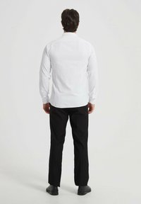 Chemise blanche à manches longues avec un col classique, au design ajusté et détails de boutons, associée à un pantalon noir et des chaussures sombres.