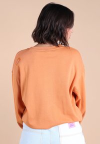 Un pull orange à manches longues avec une coupe décontractée, un col rond et une petite étiquette sur l'épaule. Associé à un jean taille haute bleu clair.