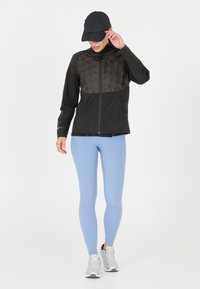 Schwarze Zip-Jacke mit einem strukturierten Oberteil, kombiniert mit hellblauen Leggings und grauen Sneakers. Das Model trägt eine schwarze Kappe.