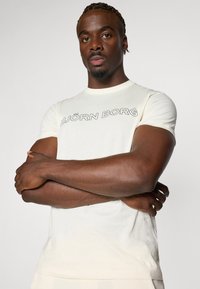 Cremfarbenes T-Shirt mit kurzen Ärmeln aus weichem Stoff, mit "BJÖRN BORG" in fetten schwarzen Buchstaben auf der Brust bedruckt.