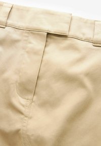Gros plan sur la taille de pantalons beiges avec passants de ceinture et panneau de braguette cousu en détail.