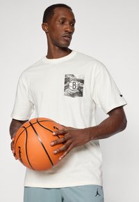 Homme portant un t-shirt blanc tenant un ballon de basket orange, le t-shirt a une poche graphique en noir et blanc et le logo Nike sur la manche.