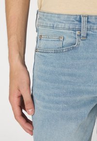 Vaqueros de mezclilla azul claro con un corte clásico, que presentan un bolsillo frontal y herrajes metálicos. La tela muestra una sutil textura desgastada.