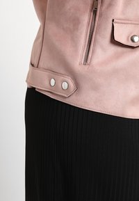 Różowa zamszowa kurtka z zapięciem na zamek błyskawiczny i bocznymi kieszeniami, wyposażona w zatrzaski. Widoczne są kontrastujące, czarne spodnie w pionowe paski.