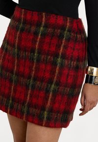 Sarkans rūtainais miniskrējs, izgatavots no pūkaina auduma ar dzelteniem un melniem akcentiem; tam ir taisns griezums un redzamās šuves gar malām.