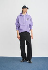 Violeta kapuce svārki ar priekšējo kabatu, saites un logo; apvienoti ar melniem platiem biksēm un melnām sporta apaviem. Zils paklāja fons.