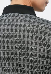 Polo com padrão a preto e branco apresentando um design geométrico, gola preta sólida e tecido texturizado.