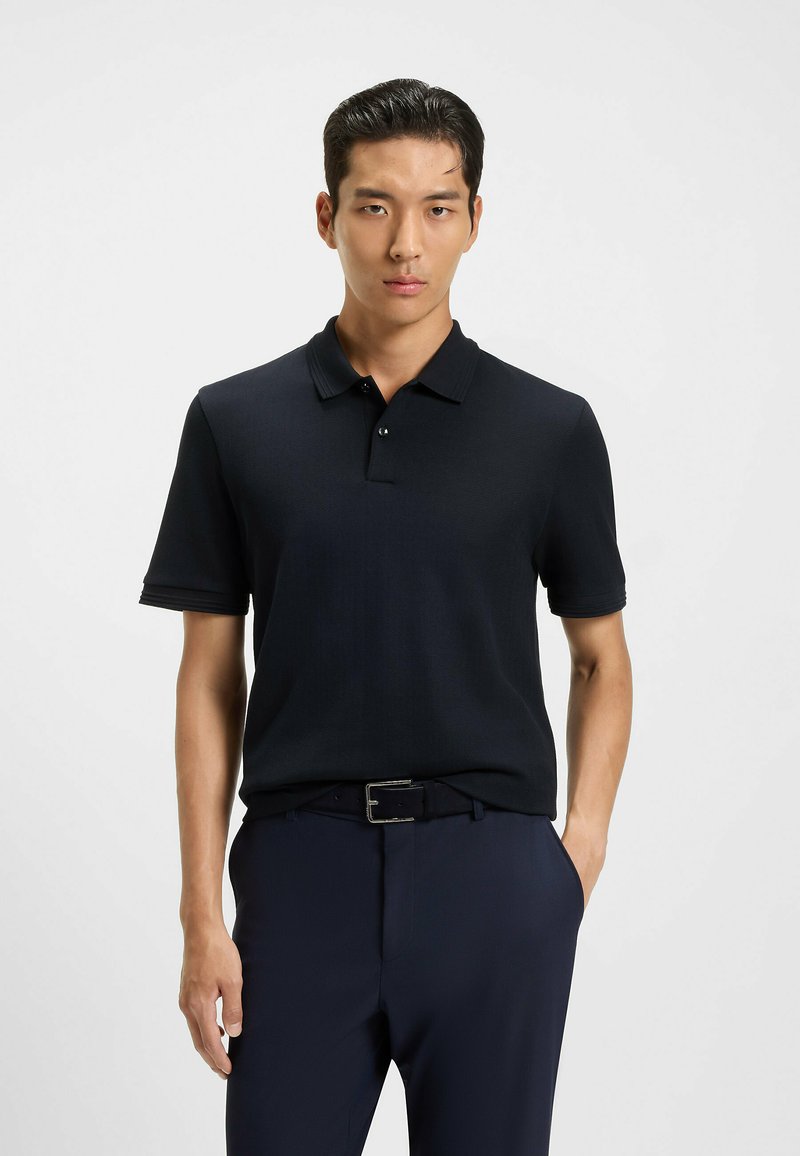 Marinblå pikétröja med kort ärm, ribbad krage och plaggstängning med tre knappar. Bärs med mörka byxor och ett enkelt svart bälte.