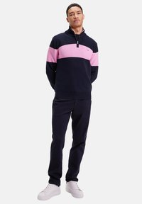 Pull-over zippé en tissu tricoté rayé bleu marine et rose, associé à un pantalon foncé et des baskets blanches, avec un logo sur le devant et des poignets côtelés.