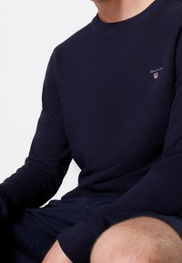 Homme portant un sweat-shirt à manches longues bleu marine avec un petit logo GANT sur la poitrine gauche, assorti à un short bleu marine.