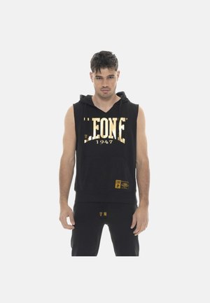 Junger Mann trägt einen ärmellosen schwarzen Hoodie mit goldener Aufschrift "LEONE 1947“ und steht aufrecht vor einem schlichten weißen Hintergrund.