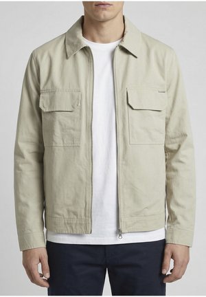 Hombre con chaqueta beige con cremallera y dos bolsillos con solapa en el pecho, sobre una camisa blanca y pantalones oscuros, de pie frente a un fondo liso.