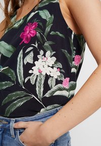 Top senza maniche con sfondo nero, decorato con motivi floreali rosa e bianchi e foglie verdi. Tessuto leggero che si adagia dolcemente.