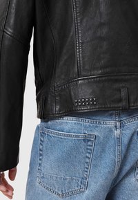 Schwarze Lederjacke mit strukturierter Oberfläche, genietetem Detail am hinteren Saum, kombiniert mit hellblauen Jeans.