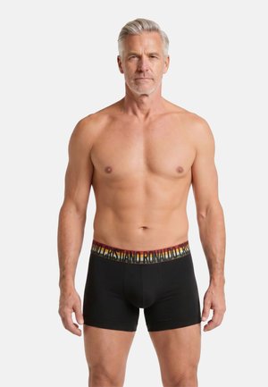 Mann mittleren Alters mit grauem Haar, der schwarze Boxershorts mit einem mehrfarbigen Bund trägt, auf denen der Text "Cristiano Ronaldo" steht, stehend vor weißem Hintergrund.