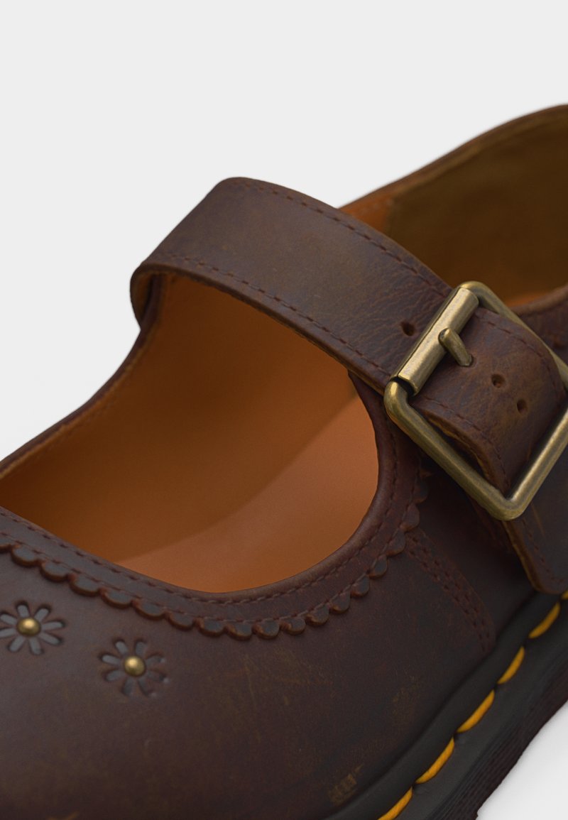 Brown leather Mary Jane shoe with scalloped edges, floral perforations, brass buckle strap, and yellow stitched sole.