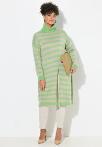Abito lungo a maglia a righe in verde chiaro e beige, con scollo alto, spacchi laterali e texture a maglia; abbinato a pantaloni beige e tacchi.