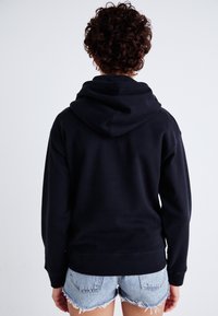 Felpa nera con cappuccio realizzata in tessuto morbido, caratterizzata da una vestibilità comoda e polsini a costine. Abbinata a pantaloni corti in denim strappato.