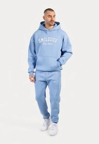 Jasnoniebieska bluza z kapturem i dopasowane spodnie dresowe wykonane z miękkiego materiału. Bluza ma kieszeń z przodu oraz biały napis z logo na bluzie i spodniach. Białe trampki.
