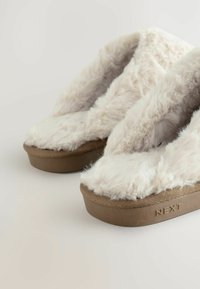Vloerbedekking beige pantoffels met een zachte kunstbont bovenkant, een afgeronde voorkant en een dikke bruine rubberen zool met een reliëf van het "NEXT"-merk.