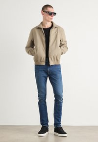 Beige Bomberjacke, schwarzes T-Shirt, blaue Jeans und schwarze Sneaker. Kurze Haare, trägt eine dunkle Sonnenbrille, mit den Händen in den Taschen.