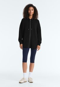 Femme portant un sweat à capuche noir zippé, des leggings bleu marine courts, des chaussettes blanches et des baskets beiges, debout sur un fond blanc uni.