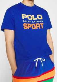 Man i blå Polo Ralph Lauren Sport t-shirt och färgglada shorts med snöre, händerna i fickorna, mot en enkel vit bakgrund.