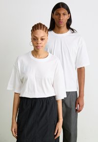 Hvite oversized t-skjorter med korte ermer, med subtil merkevare på forsiden, brukt sammen med et svart skjørt og grå bukser.