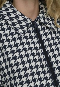 Jachetă cu fermoar cu model houndstooth în nuanțe de marină și alb, cu o guler lat și un material texturat. Fermoar negru pe partea din față.