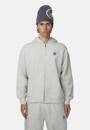 Felpa grigia con zip realizzata in materiale morbido con tasca frontale, presenta un logo blu sul petto a sinistra. Indossata con pantaloni della tuta coordinati.