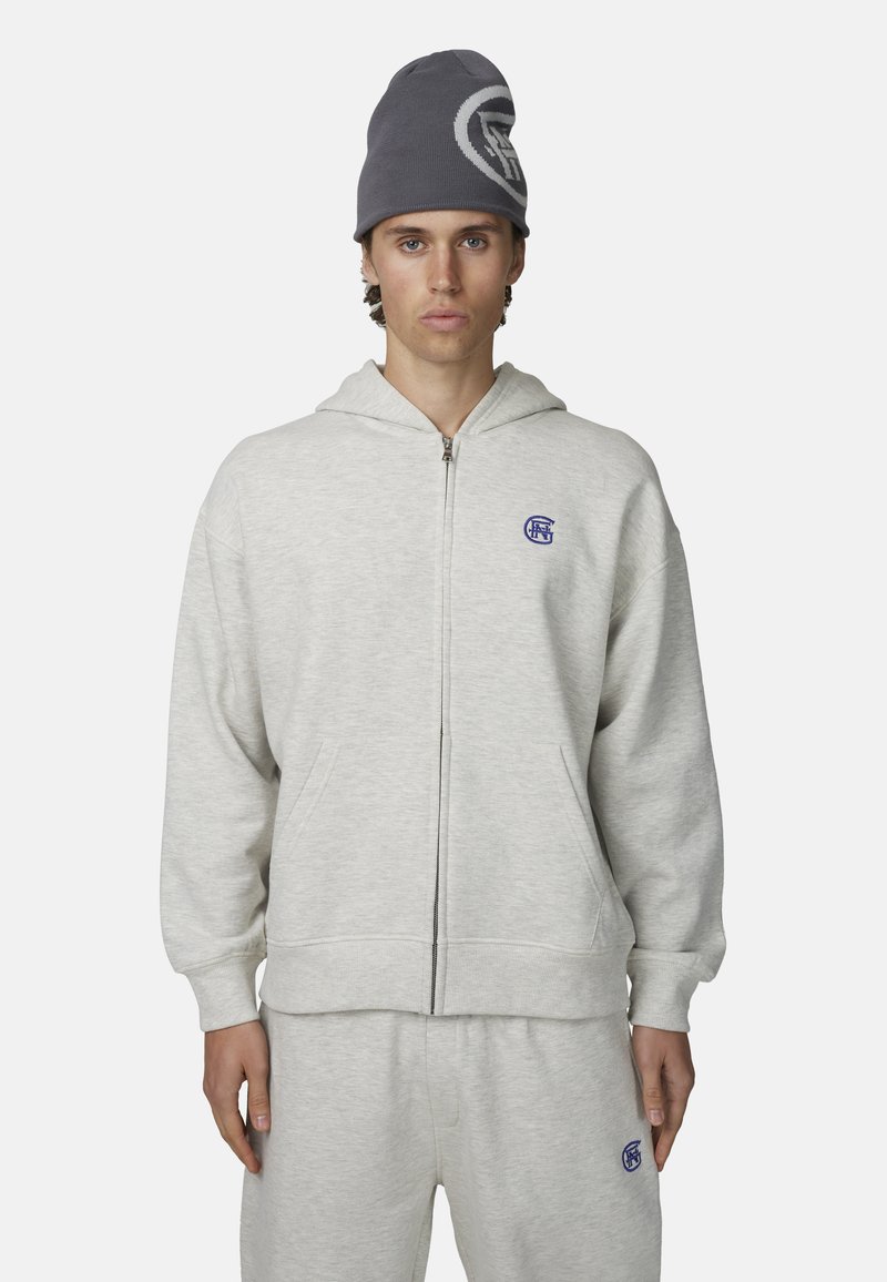 Szara bluza z zamkiem wykonana z miękkiego materiału, z kieszenią z przodu i niebieskim logo na lewej piersi. Noszona wraz z dopasowanymi spodniami dresowymi.