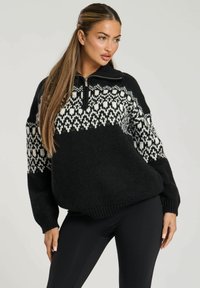 Zwarte trui met een geweven witte ontwerpen op de borst, ritssluiting bij de kraag en een dikke breistof. Gepaarde met zwarte strakke leggings.