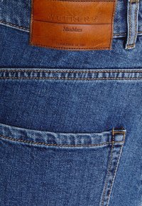 Gros plan sur la poche arrière d'un jean en denim bleu avec un patch en cuir marron portant l'étiquette "WEEKEND MaxMara Established in 1983".