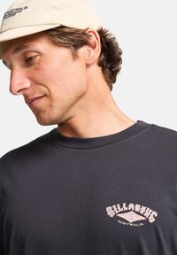 Mørkegrå T-skjorte med rund hals og korte ermer, med et rosa og hvitt "Billabong" logo-design på forsiden.