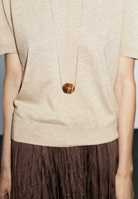 Una persona indossa un top beige e una lunga collana d'argento con un pendente marrone testurizzato, in piedi davanti a uno sfondo neutro.
