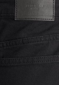 Tecido de denim preto com uma superfície texturada, apresentando uma etiqueta de couro escuro em relevo com "VERO MODA" e "DENIM" cosidas na parte superior.