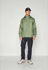 Homme se tenant debout portant une veste à capuche verte, un pantalon noir, des baskets blanches et une casquette noire devant un fond clair.