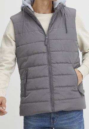 Mann trägt graue gesteppte ärmellose Weste mit Reißverschluss vorne und Reißverschlusstaschen über cremefarbenem Hoodie und blauen Jeans.