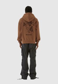 Felpa con cappuccio marrone con una grafica di una farfalla sulla schiena, abbinata a pantaloni cargo scuri con tasche laterali e polsini regolabili.