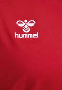 Röd bomullströja med vit broderad "hummel"-logotyp och dekorativt mönster som liknar en bi, åtsittande passform med slät textur.