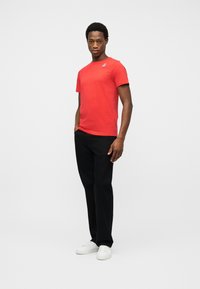 Άνδρας που στέκεται φορώντας κόκκινο κοντομάνικο πουκάμισο, μαύρο παντελόνι και λευκά αθλητικά παπούτσια σε απλό ανοιχτό φόντο.