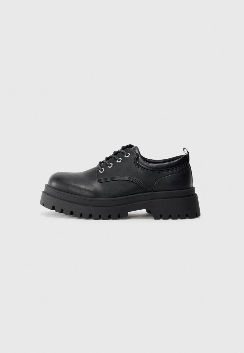 Chaussure noire en cuir à lacets avec semelle épaisse à crampons et languette de tirage, présentée sur un fond gris clair uni.