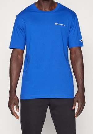 Mann trägt ein leuchtend blaues Champion-T-Shirt mit Logo auf Brust und Ärmel, kombiniert mit schwarzen Hosen, steht vor einem einfarbigen Hintergrund.