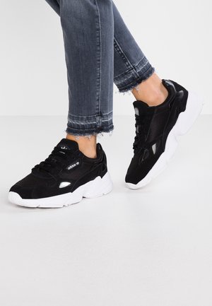 Sneaker atletiche nere con tomaia in mesh e suede, suole in gomma bianche e lacci neri. Indossate con jeans di denim strappati.