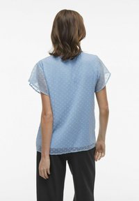 Blusa a maniche corte azzurro chiaro con un motivo a pois testurizzato, tessuto trasparente, orlo arrotondato e vestibilità ampia, abbinata a pantaloni scuri.