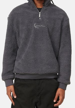 Personne portant un pull polaire texturé gris foncé avec une fermeture éclair quart de zip et un logo brodé sur la poitrine, associé à un pantalon noir.