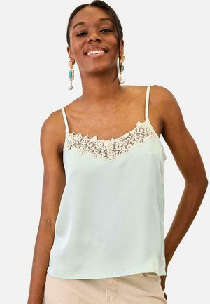 Mujer sonriente con top de satén verde pálido con ribete de encaje beige y pendientes dorados con detalles en turquesa y perla.