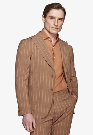 Mężczyzna w brązowym garniturze w prążki z pasującymi spodniami i pomarańczową koszulą na guziki, stoi z jedną ręką w kieszeni.