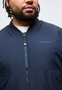 Άνδρας με πλεξούδες στα μαλλιά και γενειάδα, φορώντας μπλε σκούρο μπουφάν με φερμουάρ και το λογότυπο "Hilfiger" στο στήθος πάνω από λευκό πουκάμισο.