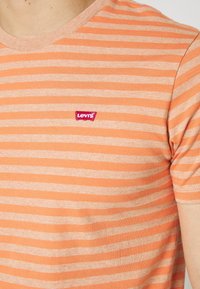 Gros plan sur un torse portant un t-shirt Levi’s rayé orange et beige avec le logo rouge Levi’s sur le côté gauche de la poitrine.